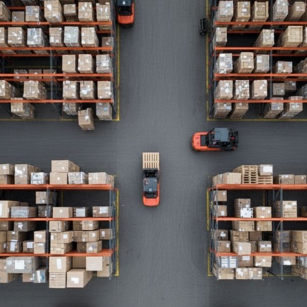 An overhead view of a warehouse shows three orange forklifts moving between tall racks stocked with numerous cardboard boxes and pallets.